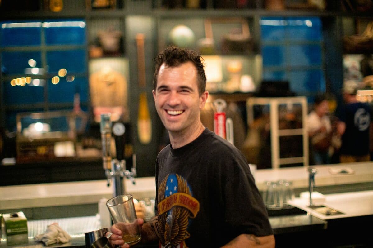 A smiling bartender holding a beer behind a bar
