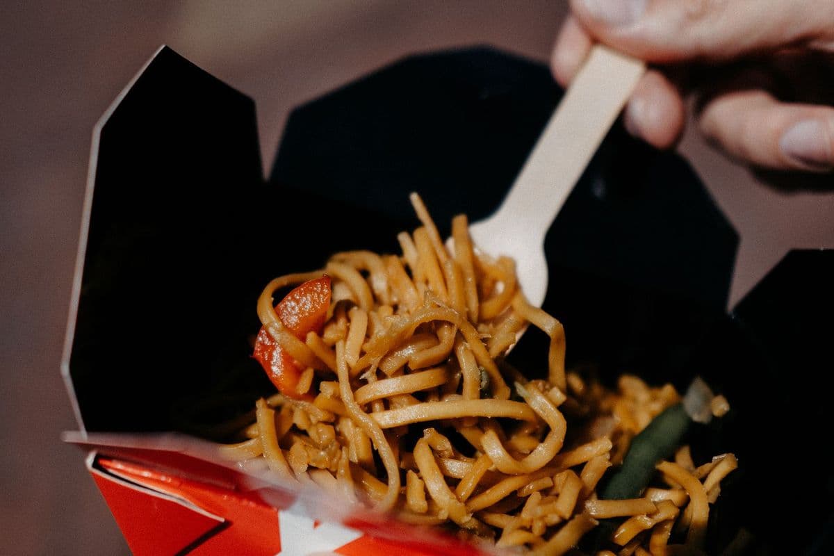 A takeaway box of noodles held open with chopsticks