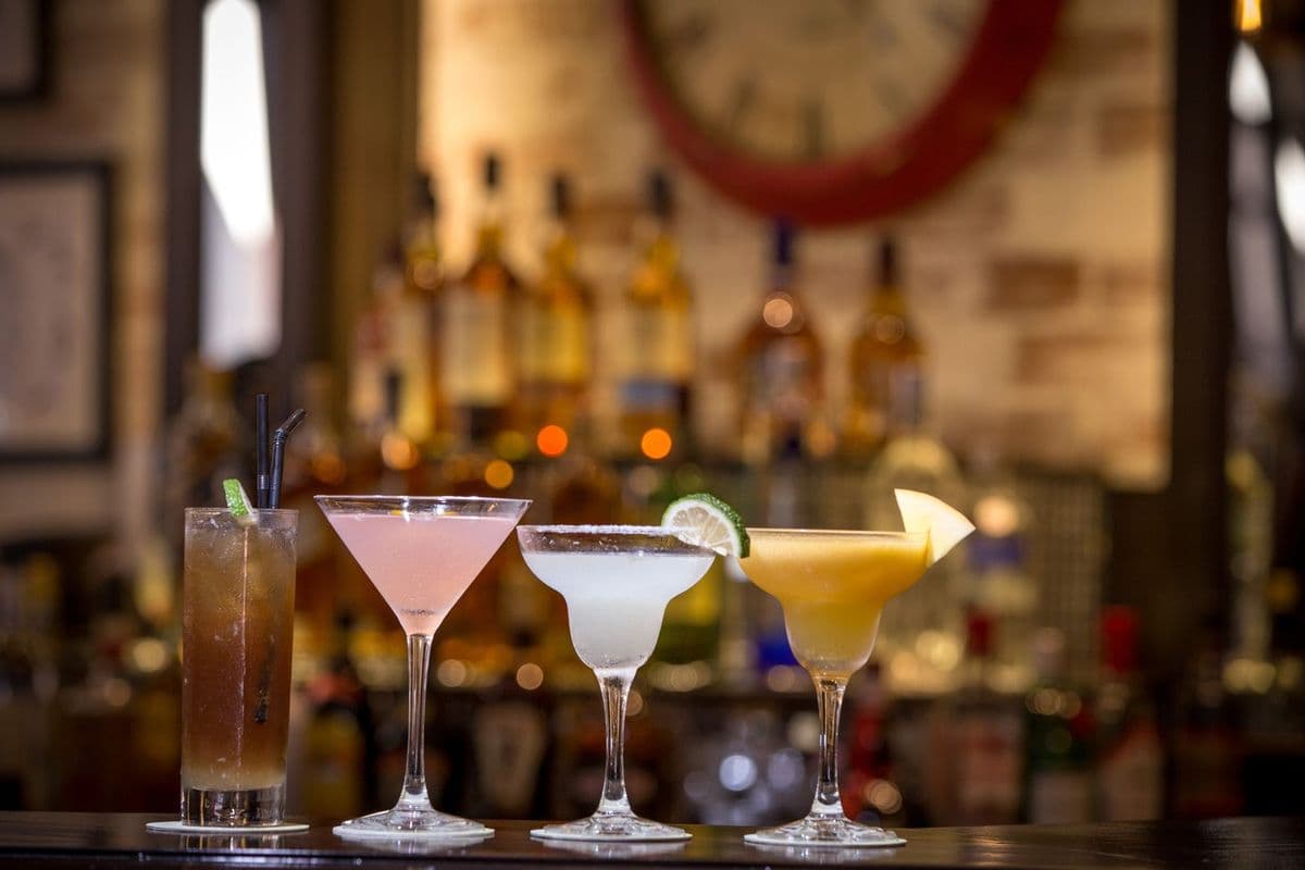 Three cocktails lined up on a bar counter with bottles behind