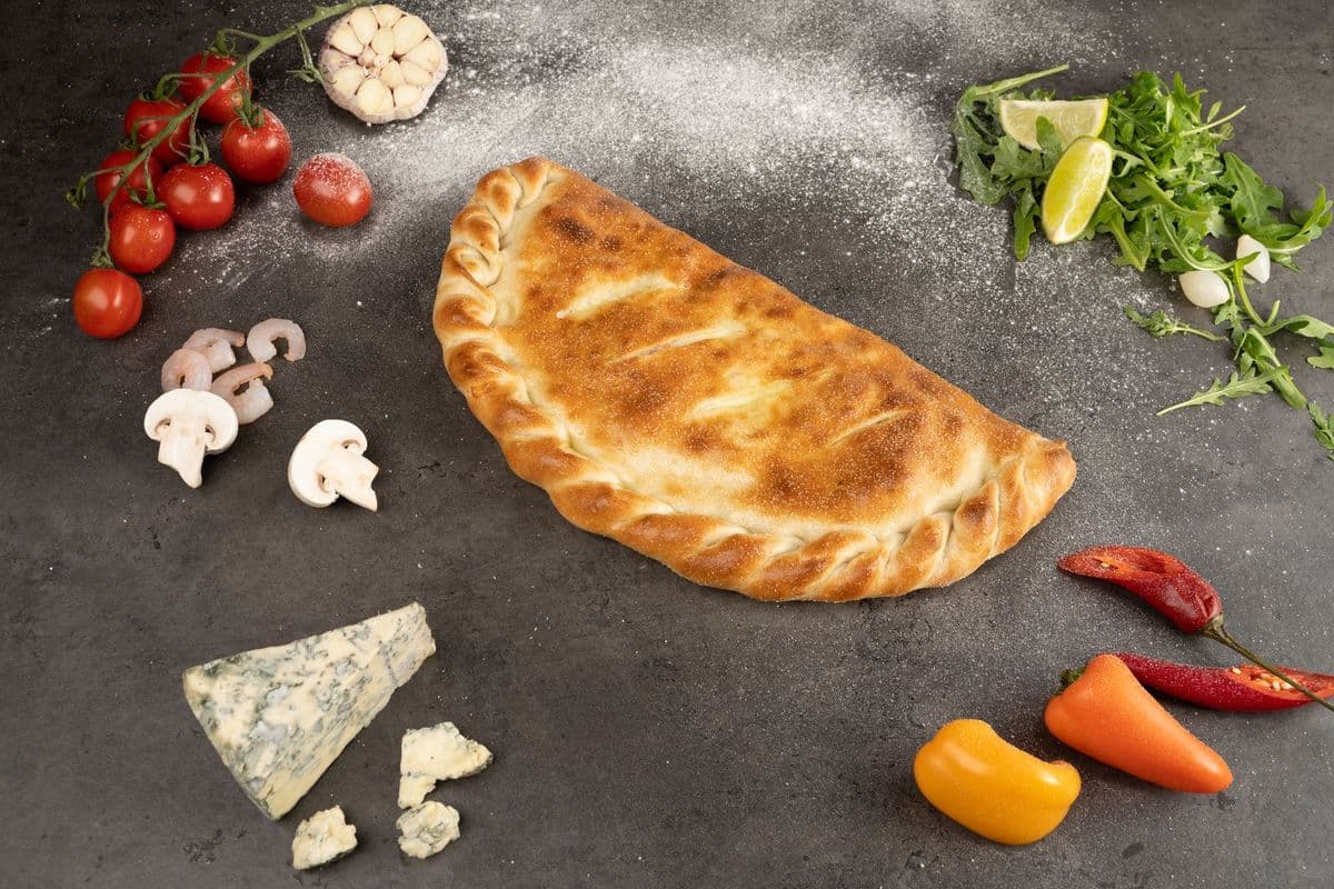 A folded calzone on a grey surface with tomatoes and herbs