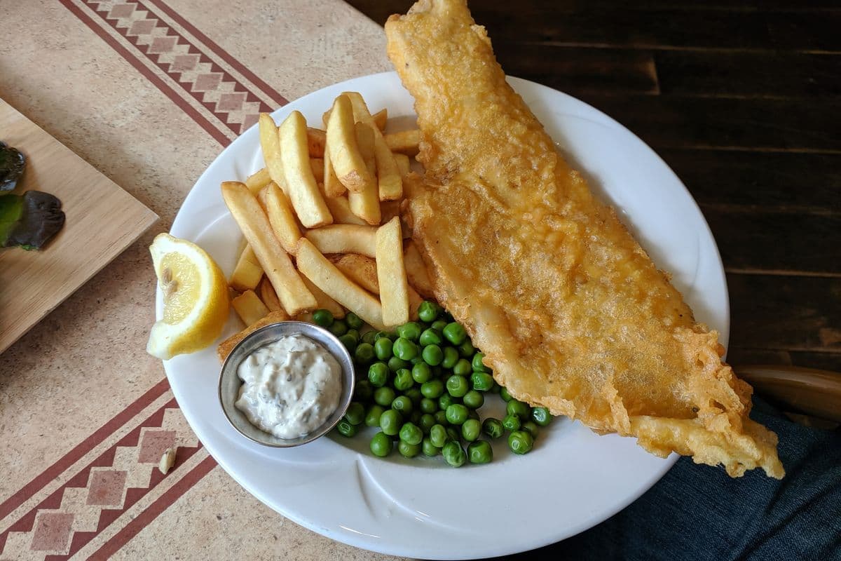 A plate of battered fish and chips with peas, a lemon wedge and tartare sauce