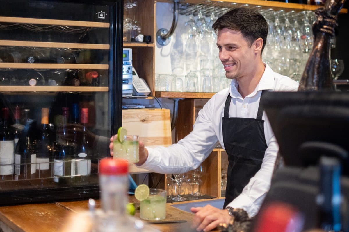 A bartender in a black apron serving a cocktail across a bar