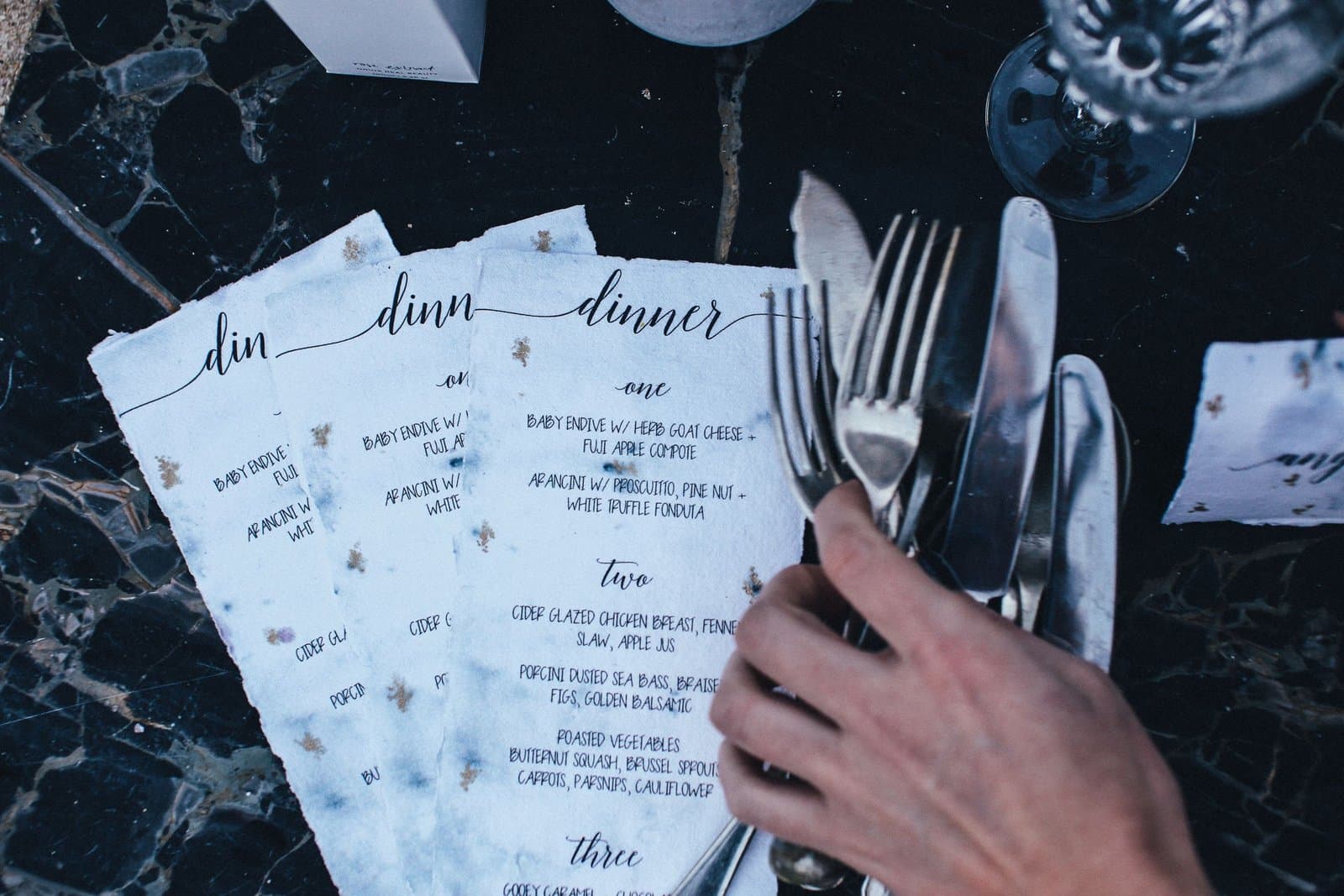Dinner menus and a hand gathering cutlery on a dark marble table