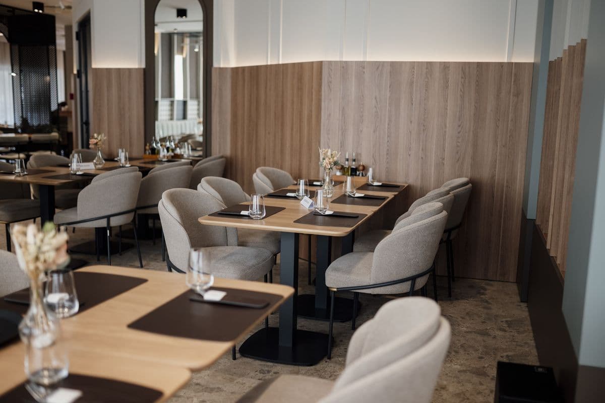 A modern restaurant dining room with wooden tables set for service and empty grey chairs