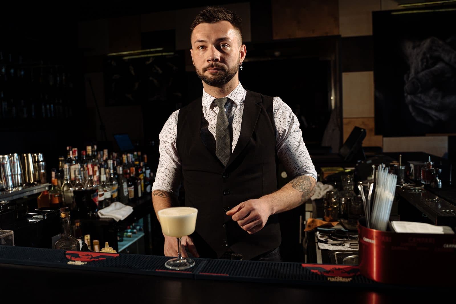 A bartender in a waistcoat standing behind a dimly lit bar