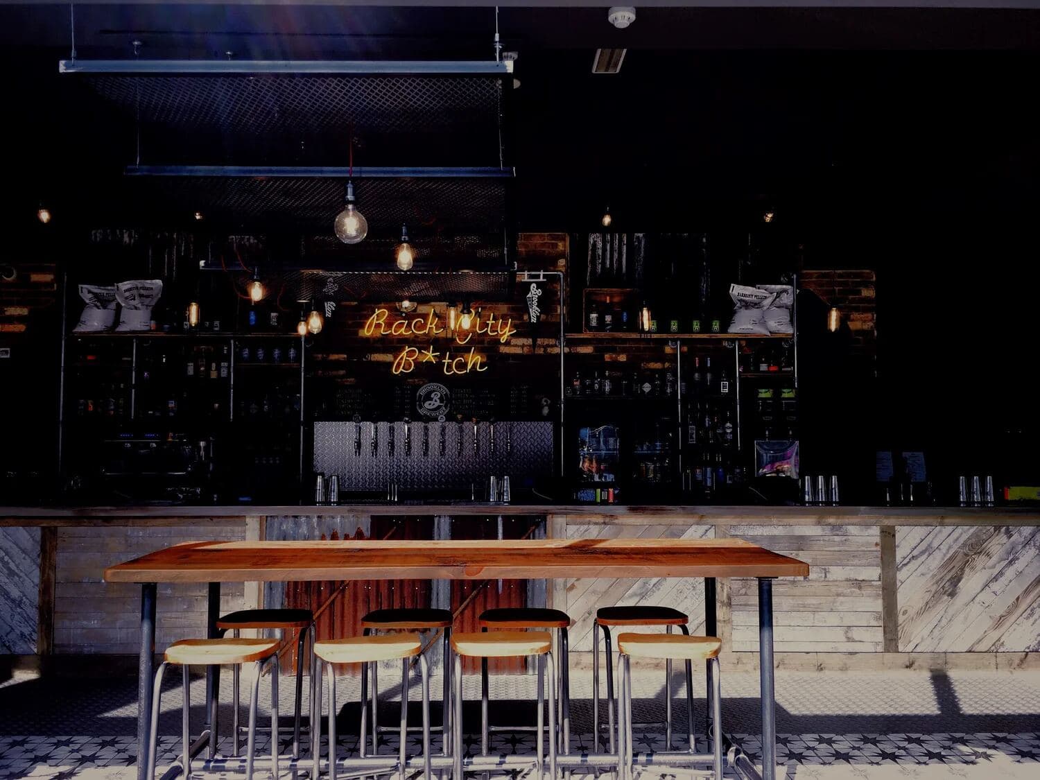 Dimly lit bar with a long wooden table, stools and a neon sign behind the back bar